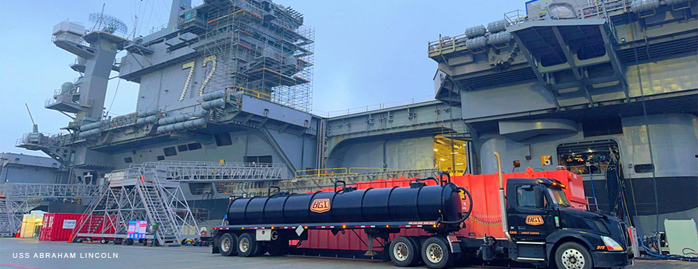 A Black Gold Industries tanker semi truck parked in front of the USS Abraham Lincoln aircraft carrier.